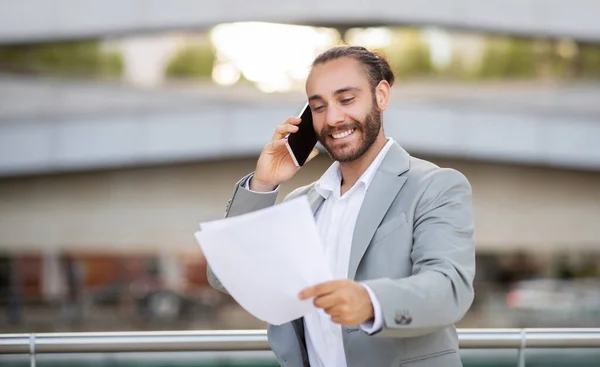 Business Lifestyle. Handsome young businessman talking on cellphone and reading documents outdoors, smiling millennial male entrepreneur having phone call with business partner, copy space