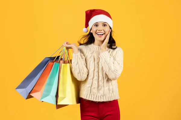 Excited young Caucasian lady buyer holding paper bags after successful Christmas sale shopping, standing over yellow studio background. New Year celebration and discounts season concept