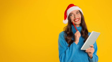 Noel süveteri ve Noel Baba şapkası giyen rüya gibi genç bayan, sarı arka planda defter ve kalem tutarak Noel dilek listesini günlüğe yazıyor. Tatil umutları ve planları. Panorama, boşluğu kopyala