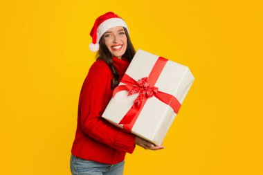 Noel hediyesi vermek. Noel Baba şapkalı gülümseyen kadın, sarı stüdyo arka planında mevcut kutuyla poz veriyor, bayram satışları ve yeni yılı kutluyor.