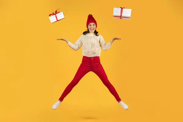 赤いニットキャップのエキサイティングなミレニアル世代の女性は 黄色いスタジオの背景でカメラに微笑んで2つのラップされたプレゼントをポーズにジャンプします 冬のバナー広告 クリスマスホリデー販売 — ストック写真