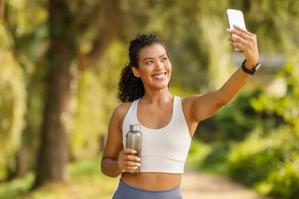 Brazilian fitness blogger lady exercising and making selfie on cellphone with water bottle in hand in park, showcasing active and connected workout routine, ideal for exercise motivation
