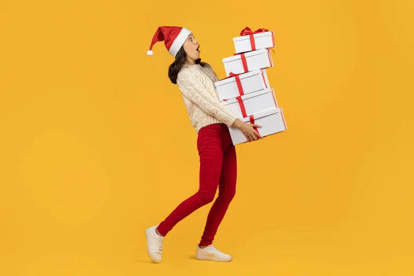 Christmas Gifts Delivery. Lady joyfully carrying stack of Xmas presents boxes on yellow studio background, wearing Santa Claus hat, representing spirit of gift giving on holiday season