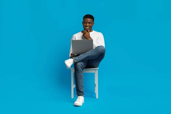 Happy curious handsome young black man in stylish outfit sitting on chair, using pc laptop on blue background, working online, have online course, copy space