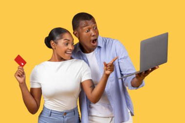 Heyecanlı siyah eşler dizüstü bilgisayarı ve kredi kartını tutuyor, kadın coşkuyla ekranı işaret ediyor, parlak sarı arka plana karşı, sezonluk satışların coşkulu havasını yakalıyor ve