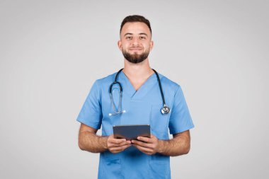 Mavi ceketli Avrupalı genç erkek doktor, elinde neşeyle dijital tablet tutuyor ve kameraya gülümsüyor, modern sağlık hizmetlerini simgeliyor, gri zemine karşı.