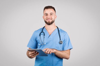 Mavi ceketli ve steteskoplu genç Avrupalı erkek doktor, dijital tablet kullanıyor, sade gri arka plana karşı online tıbbi konsültasyonu sembolize ediyor.