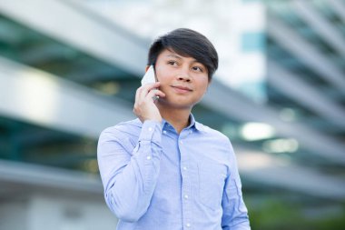 Genç Koreli ofis çalışanı akıllı telefondan iş görüşmesi yapıyor, şehir merkezinin arka planında duruyor. Şehirde telefon görüşmesi yapan başarılı Asyalı iş adamının portresi.