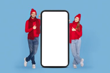 Harika bir uygulama. Örgü ören şapkalı neşeli genç çift beyaz ekranlı, akıllı telefonun yanında dikiliyor ve başparmaklarını kaldırıyor, mutlu adam ve kadın Noel reklamları sunuyor,