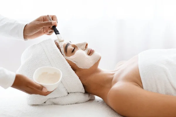 Attractive young indian woman receiving skin treatment at spa center. Professional cosmetologist doctor applying detox clay mask on beautiful eastern lady face, closeup shot with copy space