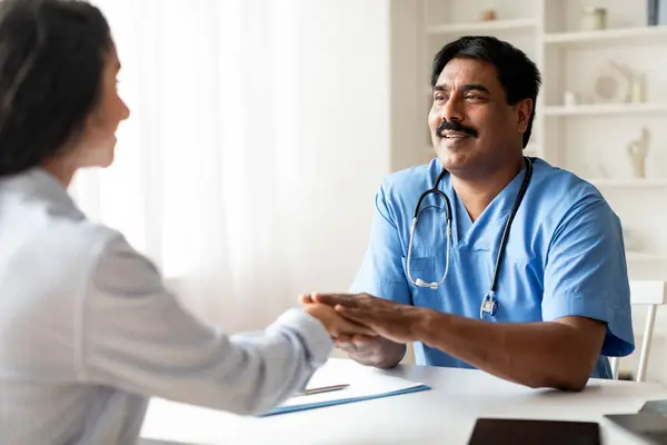 Smiling mature indian doctor man holding hand of female patient during meeting in office, smiling therapist male greeting young woman at his clinic, welcoming or greeting with successful recovery