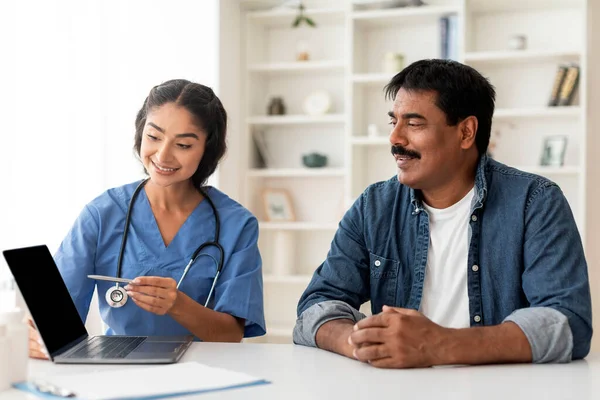 Smiling Indian Doctor Woman Showing Blank Laptop Screen To Mature Male Patient During Appointment In Clinic, Young Female Physician Discussing Treatment Process With Happy Middle Aged Man