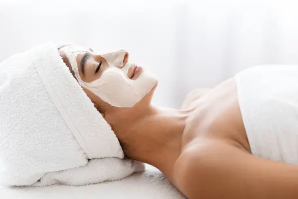 Face mask, spa beauty treatment. Side view of joyful young indian woman with white facial clay mask on her face resting on table at spa salon, skincare concept