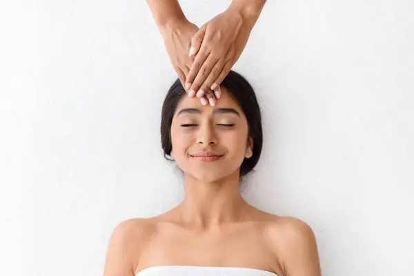 Top view of masseuse hands making relaxing massage for young beautiful indian lady, rubbing her forehead. Smiling woman getting healing head massage at modern luxury spa in romantic atmosphere