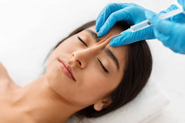 Beautician hands making hyaluronic acid injection for glabella facial rejuvenation procedure, young indian woman getting cosmetic injection between eyebrows at beauty center, top view
