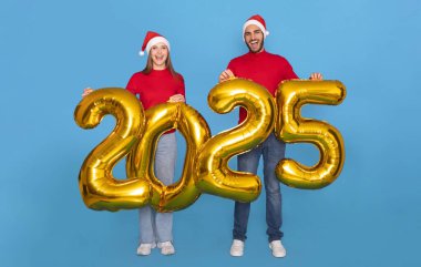 Noel Baba şapkalı genç çift, ellerinde altın 2025 balon, kameraya gülümsüyor, mutlu bir adam ve kadının mavi arka planda eğlenip, yeni yıl kutlamalarını yapıyorlar.