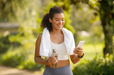 Dışarıda su şişesi ve telefon tutan genç sporcu kadın, motivasyonlu ve sağlıklı bir yaşam tarzı sergiliyor, uygulamalarda egzersiz ipuçları arıyor. Sağlık ve egzersiz konsepti