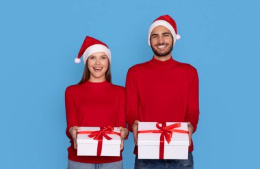 İyi tatiller. Mutlu genç çift, Noel Baba şapkalarını takıp kameraya hediye kutuları veriyorlar, Yılbaşı ve Noel 'de gülümseyen adam ve kadın, mavi arka planda tek başlarına duruyorlar.