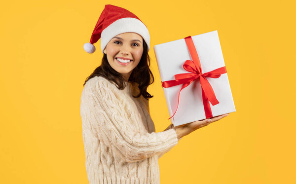 Happy lady holds New Year present with bow, posing with joyful expression in Santa hat, winter holiday shopping and gifting theme, in studio on yellow backdrop, for cheerful seasonal advertisement