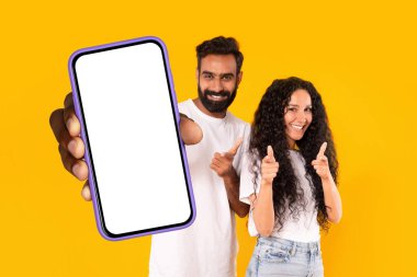 Mobil uygulama. Beyaz beyaz ekranlı, sarı arka planda duran kamerayı gösteren akıllı telefonları olan mutlu bir Arap çift. Heyecanlı adam ve kadın büyük telefon ve şablon gösteriyor