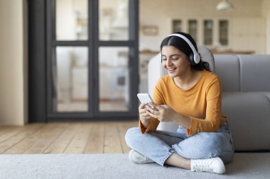 Akıllı telefonlu ve kablosuz kulaklıklı genç Hintli kadın evde dinleniyor, neşeli doğulu kadın müzik dinliyor ve oturma odasında yerde dinlenirken cep telefonuyla internette geziniyor.