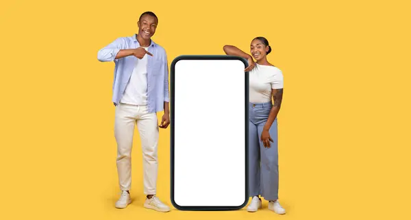 Stock image Smiling black man and woman standing next to giant smartphone, man pointing to blank screen, perfect for app or ad display, set against yellow background, panorama