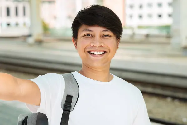 Closeup of cheerful young asian guy tourist with backpack taking selfie while waiting for his train at station, smiling at camera. Chinese man travel influencer blogger streaming while traveling