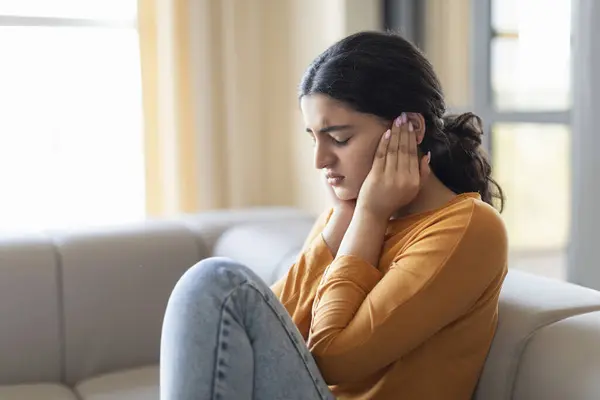 Upset young indian woman sitting on couch and covering her ears with hands, depressed eastern female expressing frustration or avoiding noise, suffering mental problems, closeup shot with copy space
