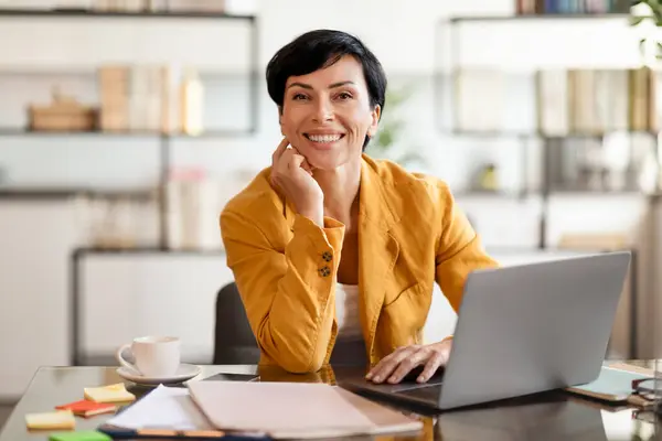 Successful entrepreneur woman posing at laptop at work desk, managing online tasks and modern technology in office. Portrait of happy middle aged businesswoman in yellow jacket at workplace