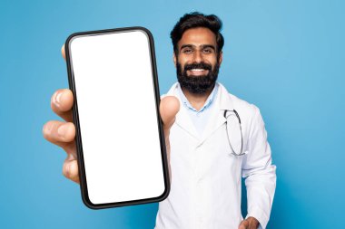 Tıbbi uygulama. Hintli doktor üniformalı, elinde boş ekranla akıllı telefon, elinde telefon olan doktor, mavi stüdyonun arka planında duruyor,