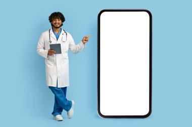 Medikal üniformalı mutlu Hintli doktor elinde dijital tabletle beyaz ekranlı büyük akıllı telefonu işaret ediyor. Çevrimiçi doktor danışmanlığı için güzel bir mobil uygulama gösteriyor.