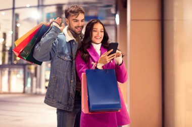 Neşeli alıcılar ellerinde alışveriş torbaları olan ve kışın mağaza dışında online satışlar için telefon başvurusu yapan çiftler, akşam alışveriş merkezlerinde mevsimlik alışverişler için mükemmel.