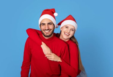 Noel Baba Şapkalı Mutlu Genç Çift 'in portresi stüdyoda mavi arka planda poz veriyor, neşeli romantik erkek ve kadın sarılıyor ve birlikte eğleniyor, Noel tatillerinin tadını çıkarıyor,