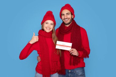 Örgü şapkalı, elinde hediye sertifikası olan gülümseyen genç bir çift kamerada başparmağını kaldırıyor, mutlu bir milenyum adamı ve kadını Noel tatili reklamları yapıyor, mavi arka planda duruyor.