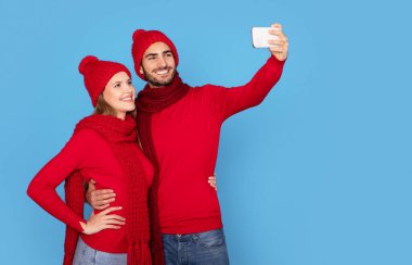 Örgü ören şapkalı mutlu çift akıllı telefondan selfie çekiyor, neşeli milenyum eşleri cep telefonlarında fotoğraf çekiyor ve birlikte eğleniyorlar, Mavi Stüdyo 'da poz veriyorlar, Uzay Kopyaları çekiyorlar.