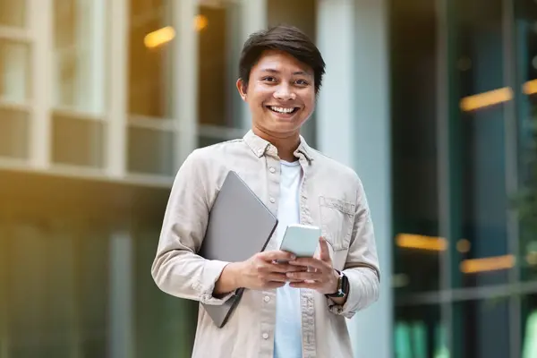 Young asian man entrepreneur wearing casual outfit with laptop computer in his hand using smartphone, standing outdoor next to office building, checking email, smiling at camera, copy space