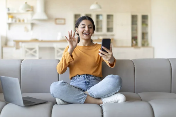 Video Call. Happy Indian Woman Teleconferencing Via Smartphone At Home, Smiling Young Eastern Female Waving Hand At Camera, Gesturing Hello To Somebody While Sitting On Couch In Living Room