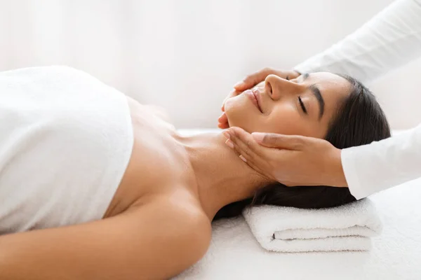 Relaxed pretty young indian woman enjoying face lifting massage in spa salon, therapist hands making skin lift procedure to relaxed brunette lady lying on table in wellness center, side view