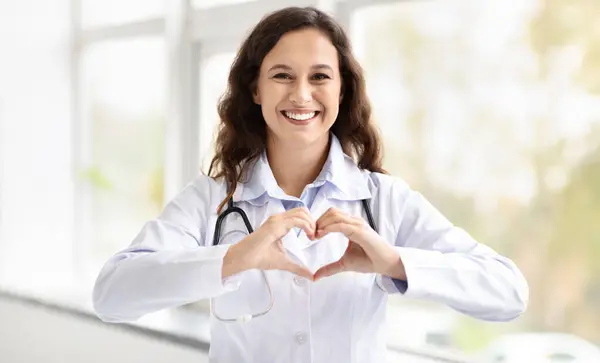 Happy millennial woman doctor showing heart shape gesture and smiling at camera, standing by window at clinic or hospital, expressing her love to patients, copy space. Medical staff