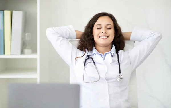 Relaxed woman doctor sitting with closed eyes and arms behind her head takes deep breath while working at office table. Stress relief for medical staff concept, copy space