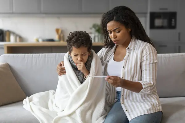 Caring Black Mom Checking Temperature Of Her Sick Son At Home, Worried Mother Holding Thermometer And Embracing Ill Little Boy, Male Kid Sitting Covered In Blanket And Coughing Into Fist