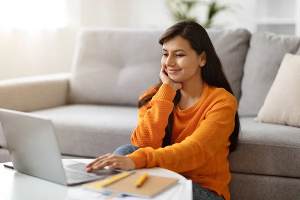 Beautiful young indian woman using laptop at home, sitting on floor next to couch in living room, websurfing or scrolling, booking traveling tour online, checking nice offer, copy space