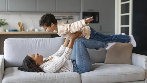 家庭游戏 快乐的黑人妈妈和小儿子在家里玩耍 躺在沙发上 像飞机一样抱着自己的男孩儿 牵着男孩的手 孩子们假装像翅膀一样张开着双手 — 图库照片