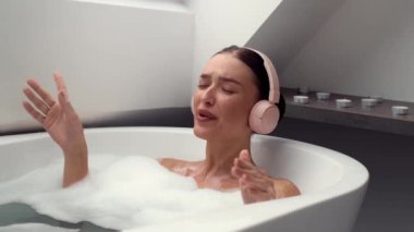 Sakin banyo atmosferinde, 4K yavaş çekim videosunda köpük banyosu yapan, kulaklık takan ve neşeyle şarkı söyleyen bir kadın görülüyor. Bu görüntüler rahatlama ve müziğin mutlu birleşmesini özetliyor..