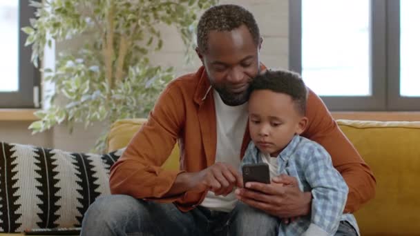 現代家族の時間 ハッピーブラックの父親と彼の息子は スマートフォンで教育アプリを探索し 自宅での生活 追跡ショット フリースペースで技術の重要性を示しています — ストック動画