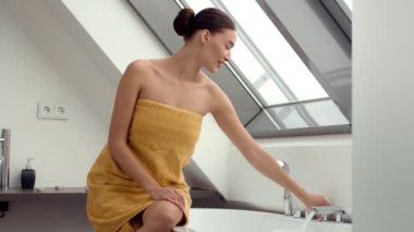 Yavaş çekim 4K videosu havluya sarılı genç bir kadını banyoyu doldurmak ve daha fazla köpük yaratmak için musluğu açarken yakalıyor. Sahne, rahatlatıcı bir banyo ayininin heyecanını ve beklentilerini tasvir ediyor..