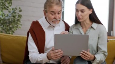 E-okuryazarlık konsepti. Yetişkin torun, büyükbabasına dizüstü bilgisayar ve internet kullanmayı öğretiyor, evdeki koltukta oturuyor, çekimleri takip ediyor, yavaş çekim, boş alan.