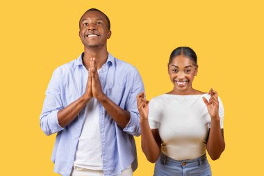 Siyahi adam ellerini sıkarak dua ediyor ve kadın parmaklarını çapraz yapıp gülümsüyor. İkisi de sarı arka planda en iyi şekilde durmayı umuyor.