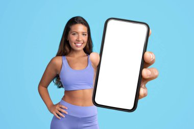 Spor salonundaki atletik bayan boş ekran modeli olan büyük bir telefon sergiliyor, egzersiz uygulamaları ve online fitness iletişimi için ideal, mavi stüdyo arka planının başında duruyor. Kolaj