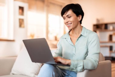 Gülümseyen orta yaşlı iş kadını dizüstü bilgisayarda çalışıyor, koltuğunda gelişigüzel oturuyor, modern oturma odasında kariyer ve konforu birleştiriyor, serbest yaşam tarzının portresi